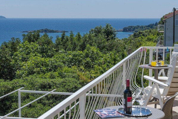 A balcony with a view of the sea and greenery, a table with a bottle and glasses, and a small table with a drink.