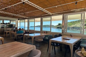 Un restaurant doté de grandes fenêtres offrant une vue sur la mer et les collines. Des tables et des chaises sont disposées pour accueillir les clients.