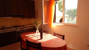 Een eettafel met een rode tafelkleed, twee wijnglazen en een fles wijn. Op tafel staat een vaas met bloemen. De kamer heeft houten kastjes en een raam met gordijnen.