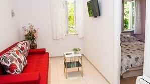 Una habitación compacta con un sofá rojo, una mesa pequeña con un florero, un televisor montado en la pared y una cama con colcha estampada.