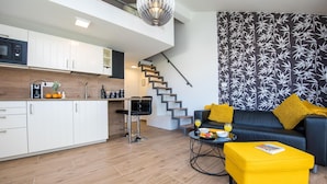 A modern living space with a kitchen, a staircase, a sofa with yellow cushions, and a coffee table with fruits and a magazine.