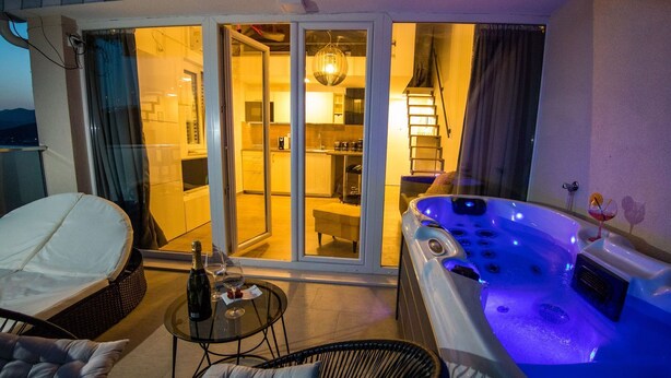 A balcony with a hot tub, wicker chairs, a glass table with a bottle and glass, and a sliding glass door leading to a kitchen.