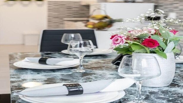 Een eettafel gedekt met witte borden, servetten en wijnglazen, met een bloevas ernaast.