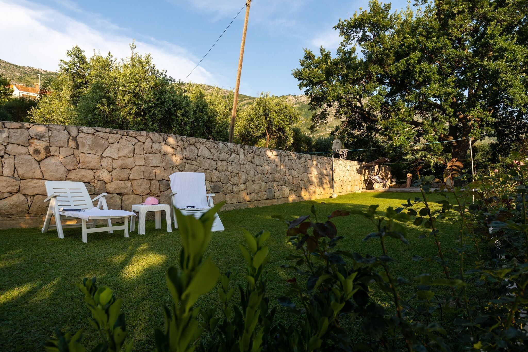 Un mur de pierres, deux fauteuils de jardin blancs et une petite table, le tout dans un jardin où l’herbe est verte et où poussent des arbres.