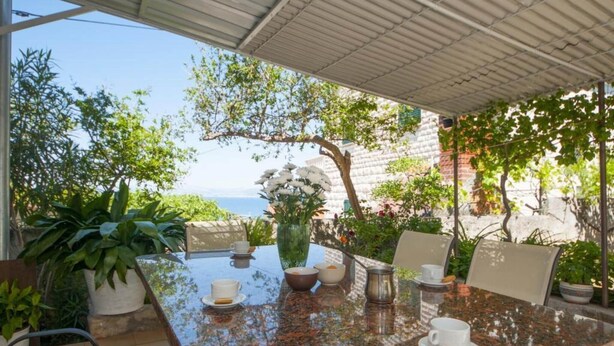 Un patio al aire libre con una mesa lista para dos personas, rodeado de macetas con plantas y vistas al mar.