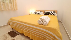 Una cama bien hecha con una colcha amarilla y almohadas a rayas blancas, una toalla enrollada sobre la cama y una lámpara de noche pequeña.