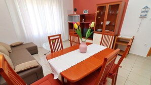 Un comedor con una mesa de madera, sillas y un jarrón con flores. Se aprecia un sofá y un aparador al fondo.
