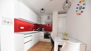 Una cocina moderna con armarios blancos, un salpicadero rojo y una zona de comedor con mesa de vidrio y sillas blancas.