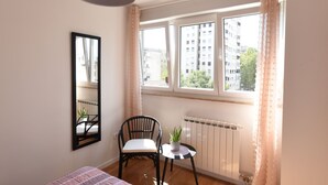 Un dormitorio con una cama, una silla, una mesita con una planta, una ventana con cortinas y un radiador.