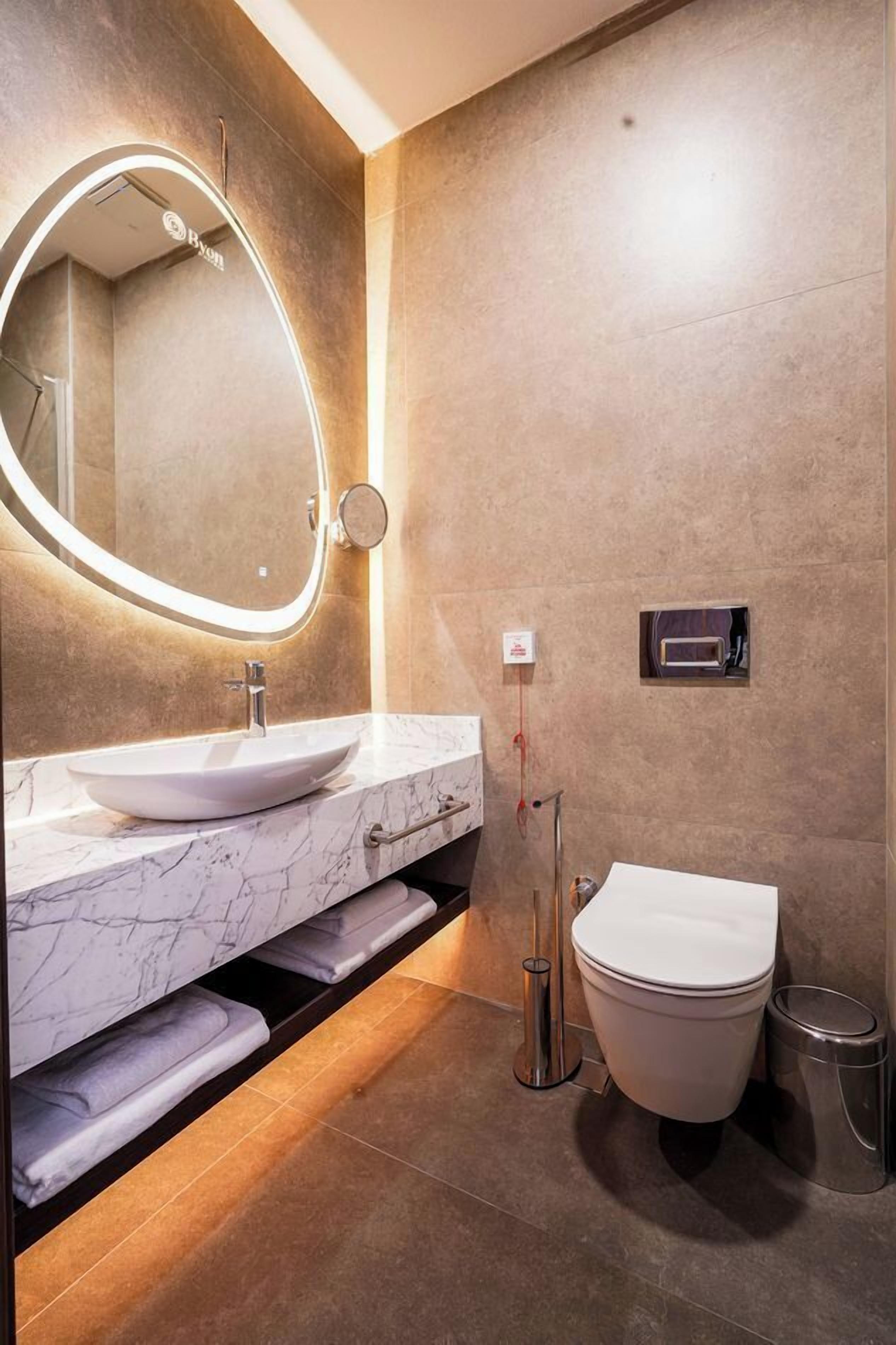 Un baño moderno con tocador de mármol, un espejo redondo con iluminación, un inodoro y un cubo de basura.