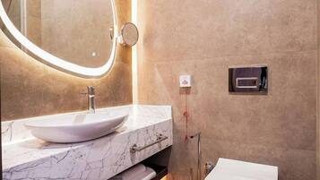 Un baño moderno con tocador de mármol, un espejo redondo con iluminación, un inodoro y un cubo de basura.