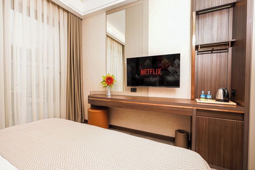 Una habitación de hotel con cama, un escritorio de madera, un televisor mostrando el logotipo de Netflix, un florero con flores y una ventana con cortinas.