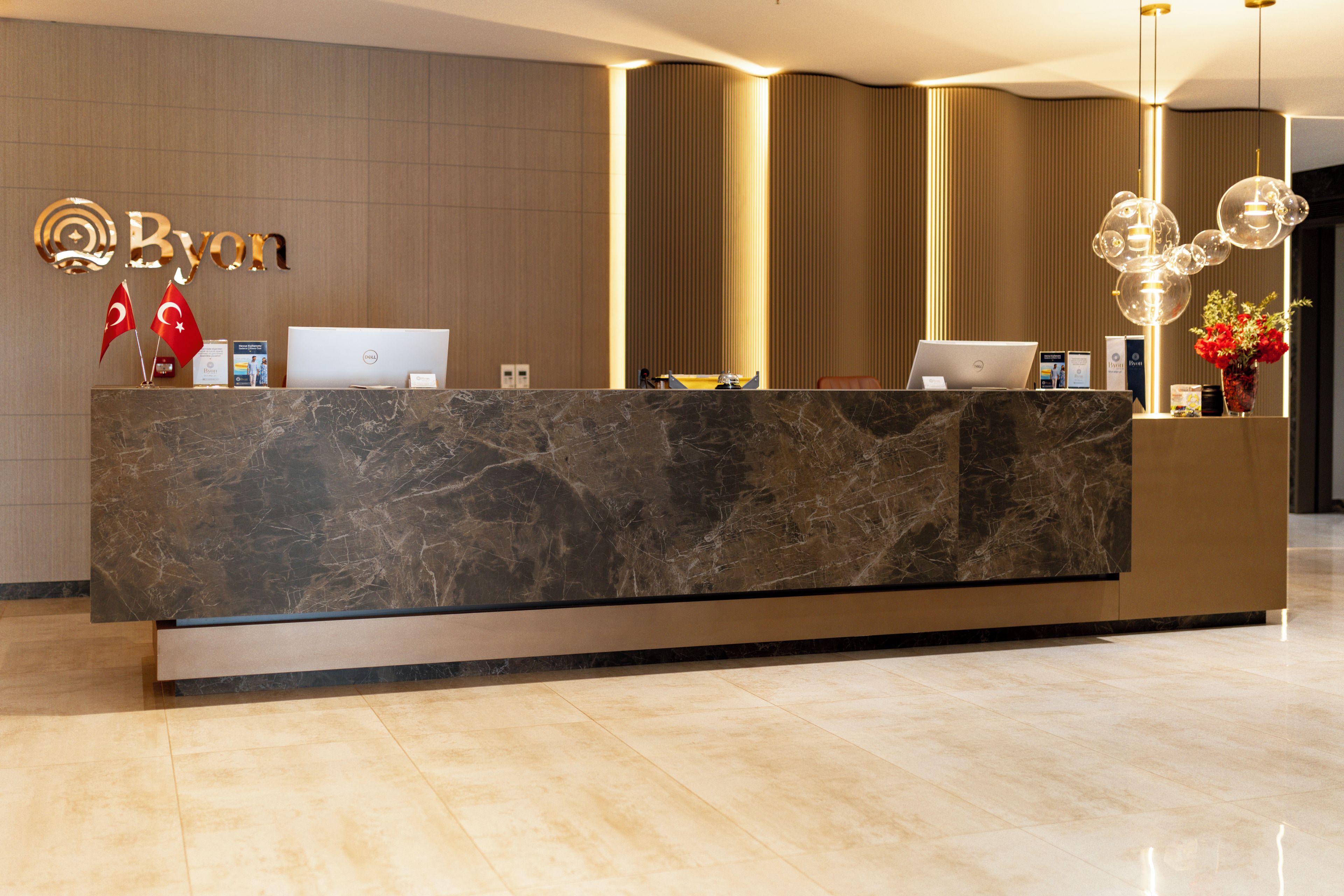 A reception desk with a marble top, two laptops, a vase with red flowers, and flags on the wall.