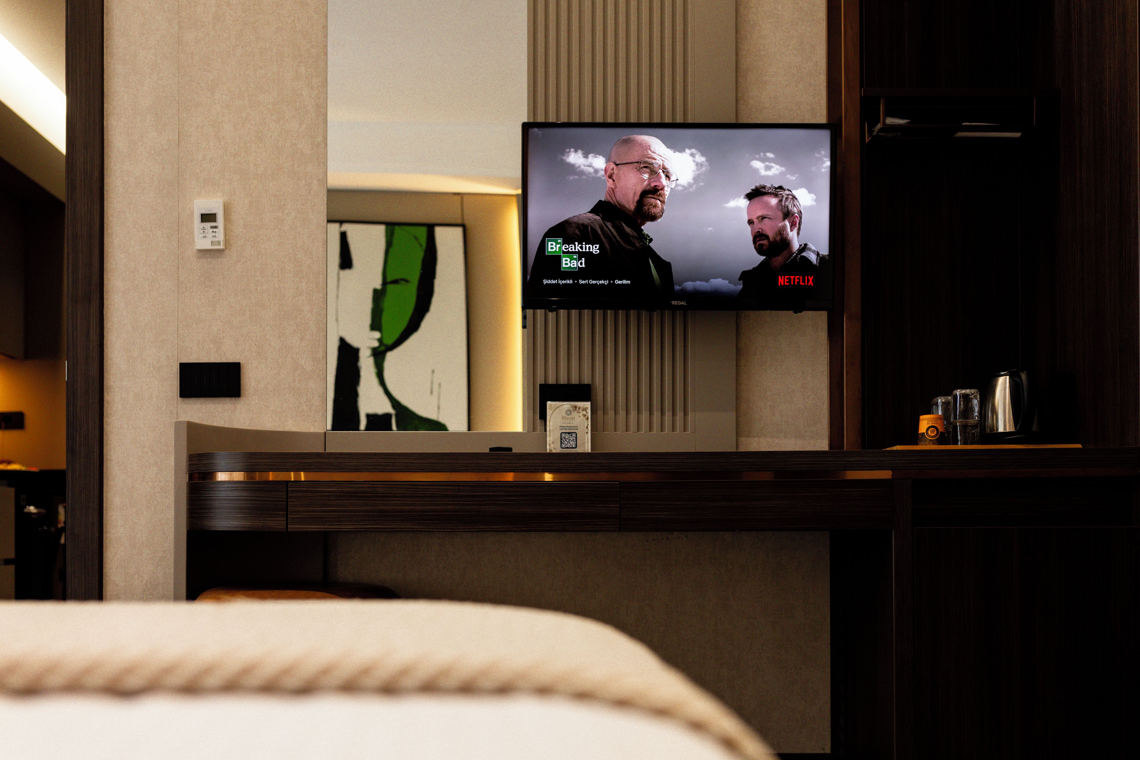 A hotel room with a television mounted on the wall, a bedside table with a coffee maker, and a framed artwork on the wall.