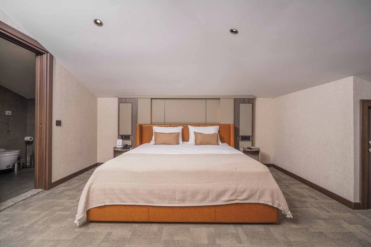 A large bed with a wooden frame and a beige patterned bedspread, flanked by nightstands with lamps, in a room with a neutral color palette and a visible bathroom door.