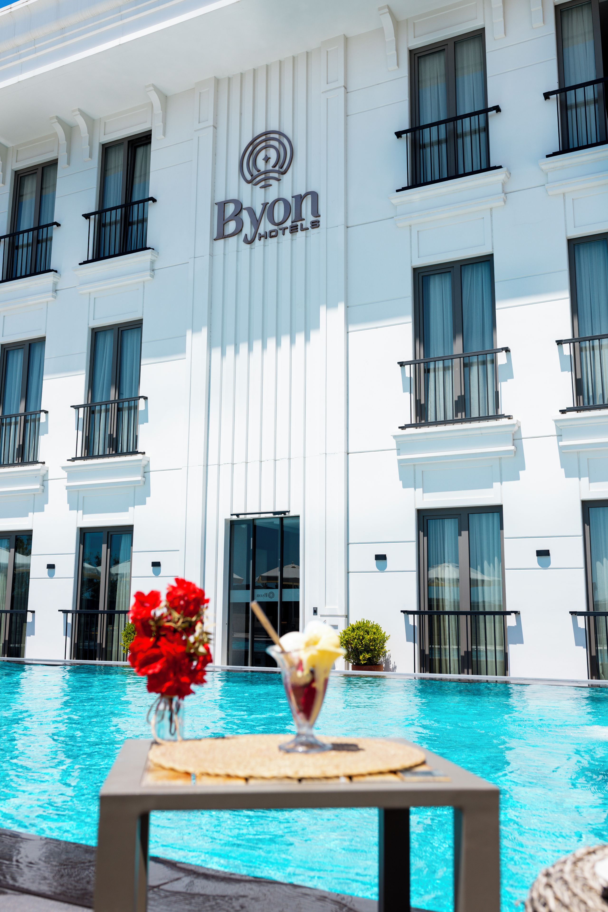 Un hotel con piscina, una mesa con una bebida y flores rojas.
