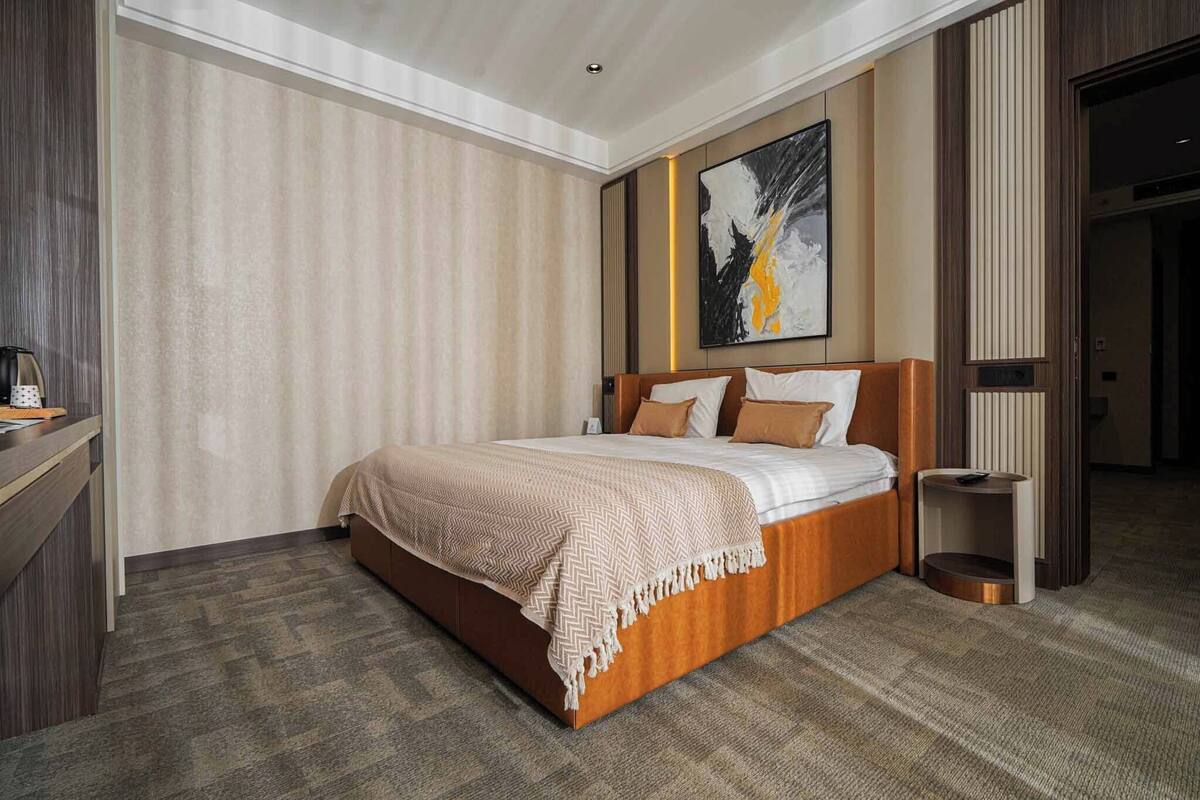 A hotel room with a large bed, a bedside table, a wall-mounted artwork, and a door leading to another room.