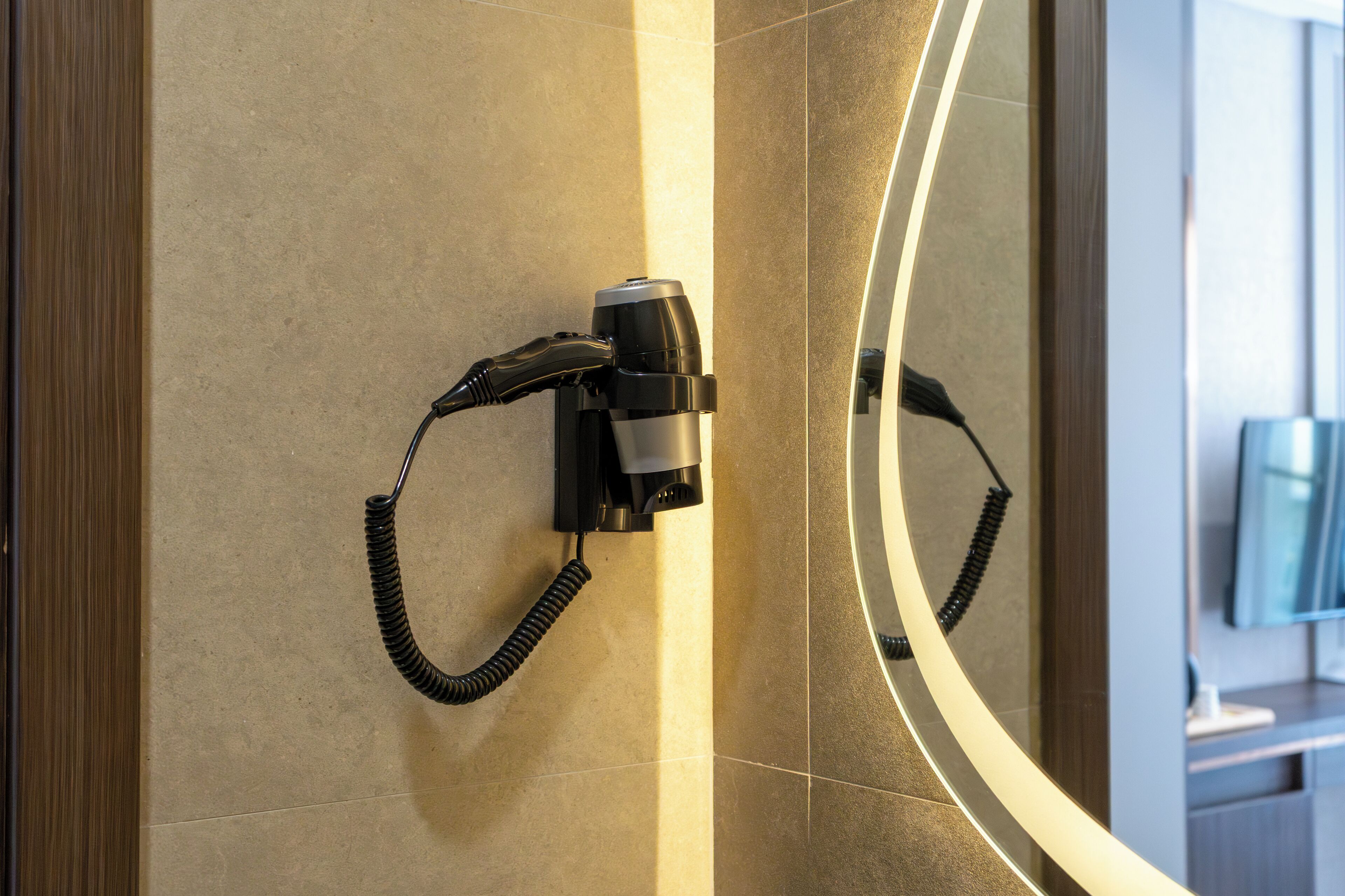 Un secador de pelo montado en la pared con un cable, un espejo que refleja parte de la habitación y un televisor al fondo.
