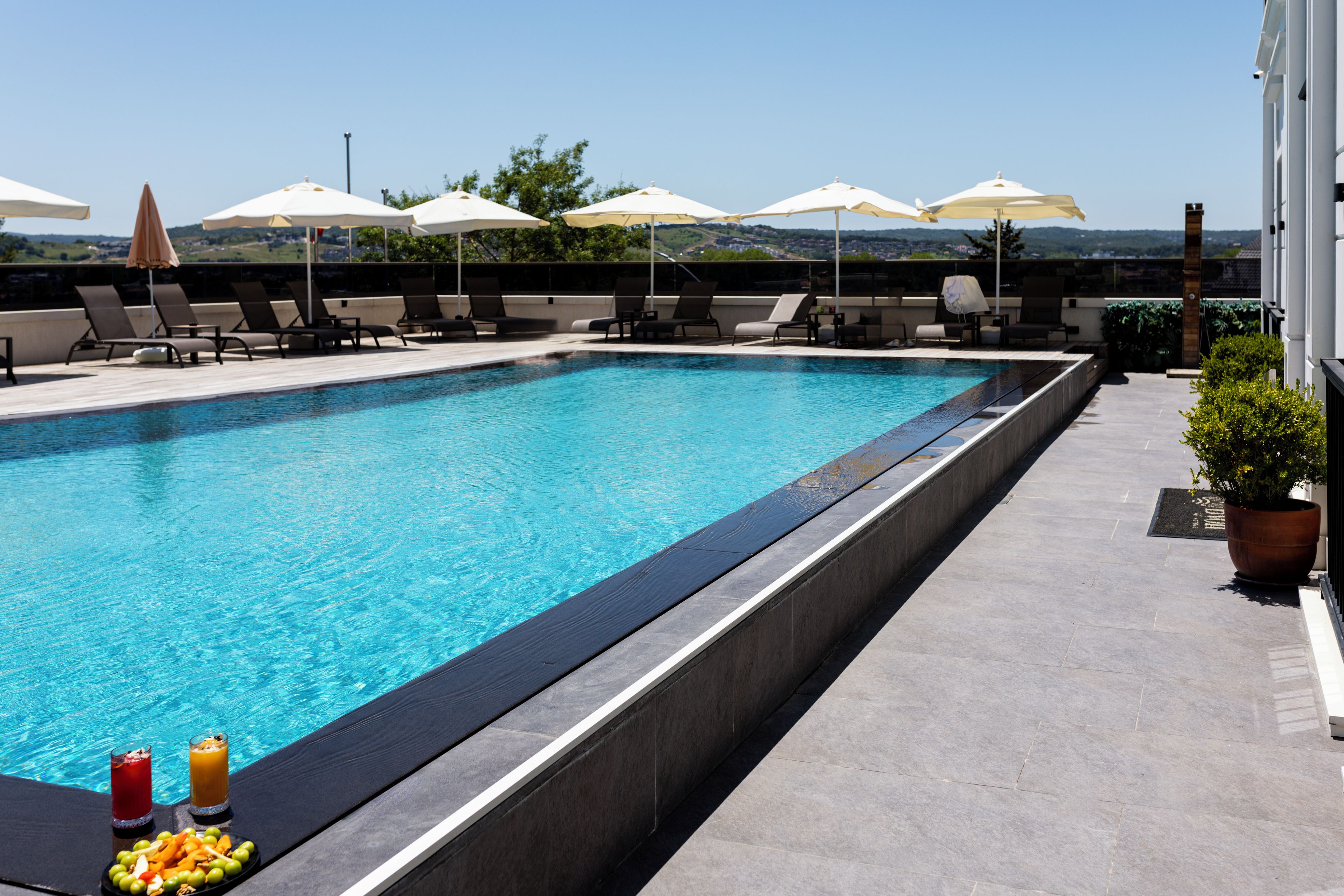 A rectangular swimming pool with a tiled edge, surrounded by lounge chairs and umbrellas, with a view of a hilly landscape in the background.