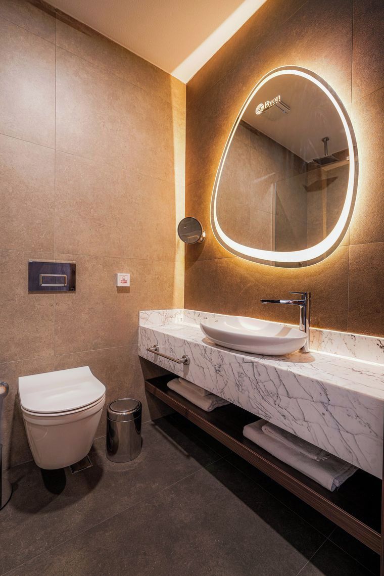 Et moderne badeværelse med et hvidt toilet, en vask med marmorbordplade og et spejl monteret på væggen med cirkulær belysning.