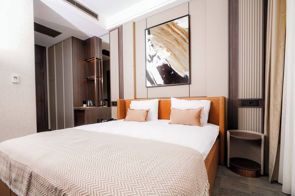 A hotel room with a large bed, a bedside table, a mirror, a painting on the wall, and a small trash bin.