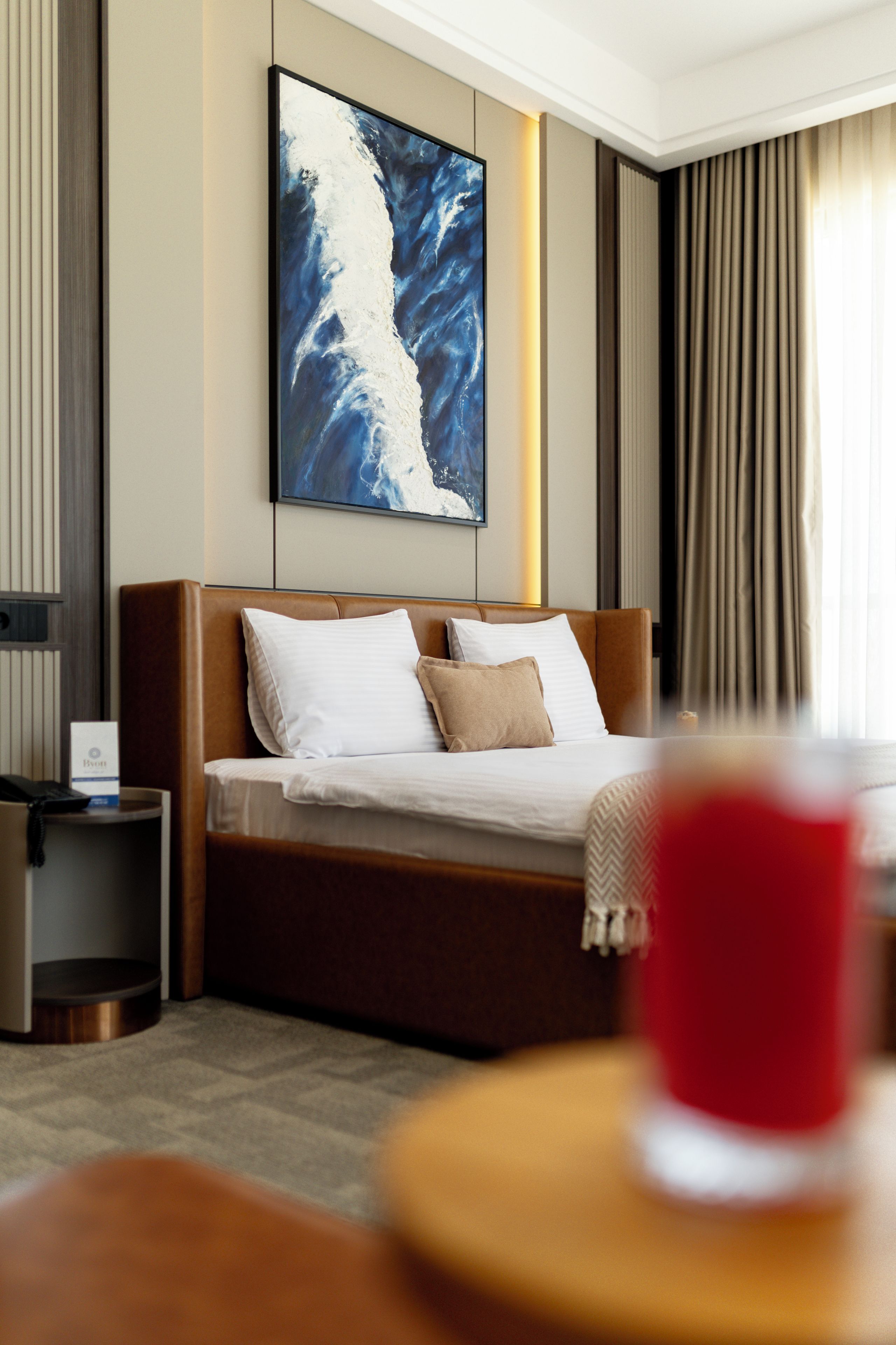A hotel room with a bed, a bedside table, a wall-mounted painting, and a window with curtains.