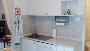 Una cocina compacta con lavamanos, lavavajillas y una pequeña encimera. Hay armarios sobre el lavamanos y una estantería con objetos decorativos.