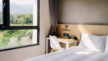 Habitación de hotel con cama, escritorio con silla, mesita de noche, ventana con vistas a zonas verdes y montañas, y cortina.