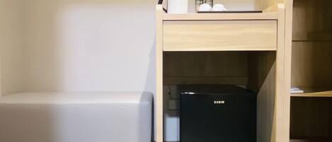 A compact room with a bed, a small black refrigerator, and a wooden desk with a cup and a bottle on top.