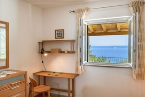 A room with a wooden desk, a chair, a small shelf with a picture, and a window with a view of the sea.