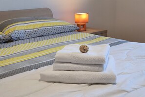 Un lit bien fait, avec une couette blanche et des serviettes pliées, une couverture à motifs et une lampe de chevet.