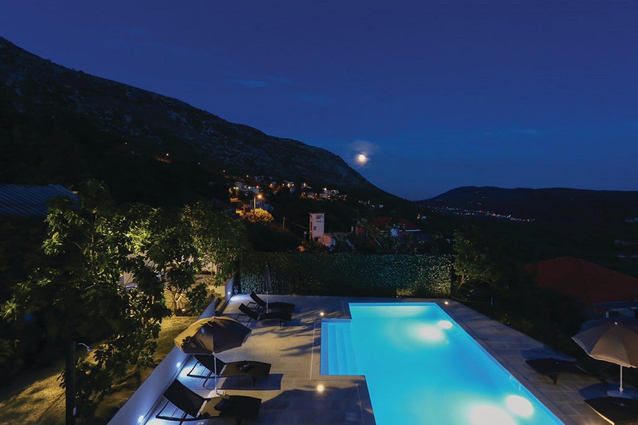 Ein Swimmingpool mit Liegestühlen und Sonnenschirmen, umgeben von viel Grün und einer Bergkulisse.