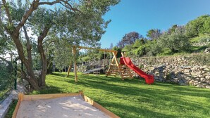 Un parque infantil con tobogán y columpios, rodeado de un muro de piedra y árboles.