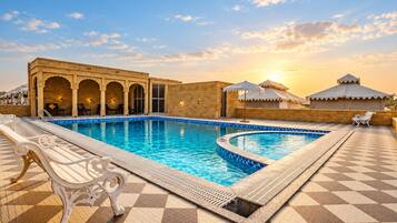 Una piscina con suelo a cuadros, rodeada por un edificio con arcos y el sol poniéndose al fondo.