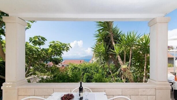 A table with a bottle and glasses, a bowl of fruit, and a view of greenery and buildings.