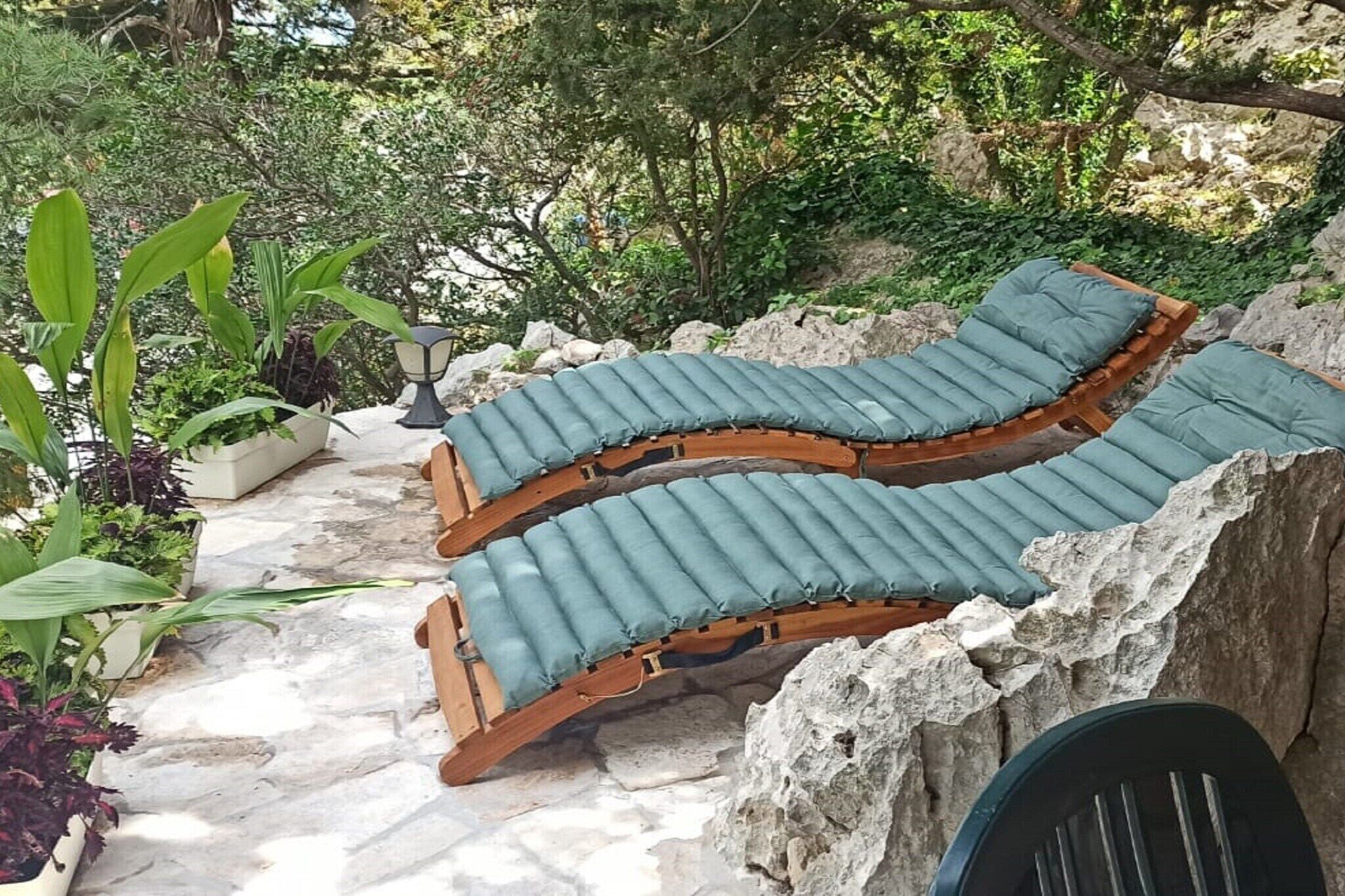 Un banco curvo de madera con cojines verdes en un jardín con suelo de piedra y plantas en macetas.