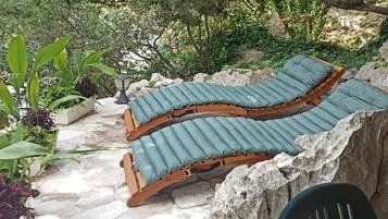 Un banco curvo de madera con cojines verdes en un jardín con suelo de piedra y plantas en macetas.