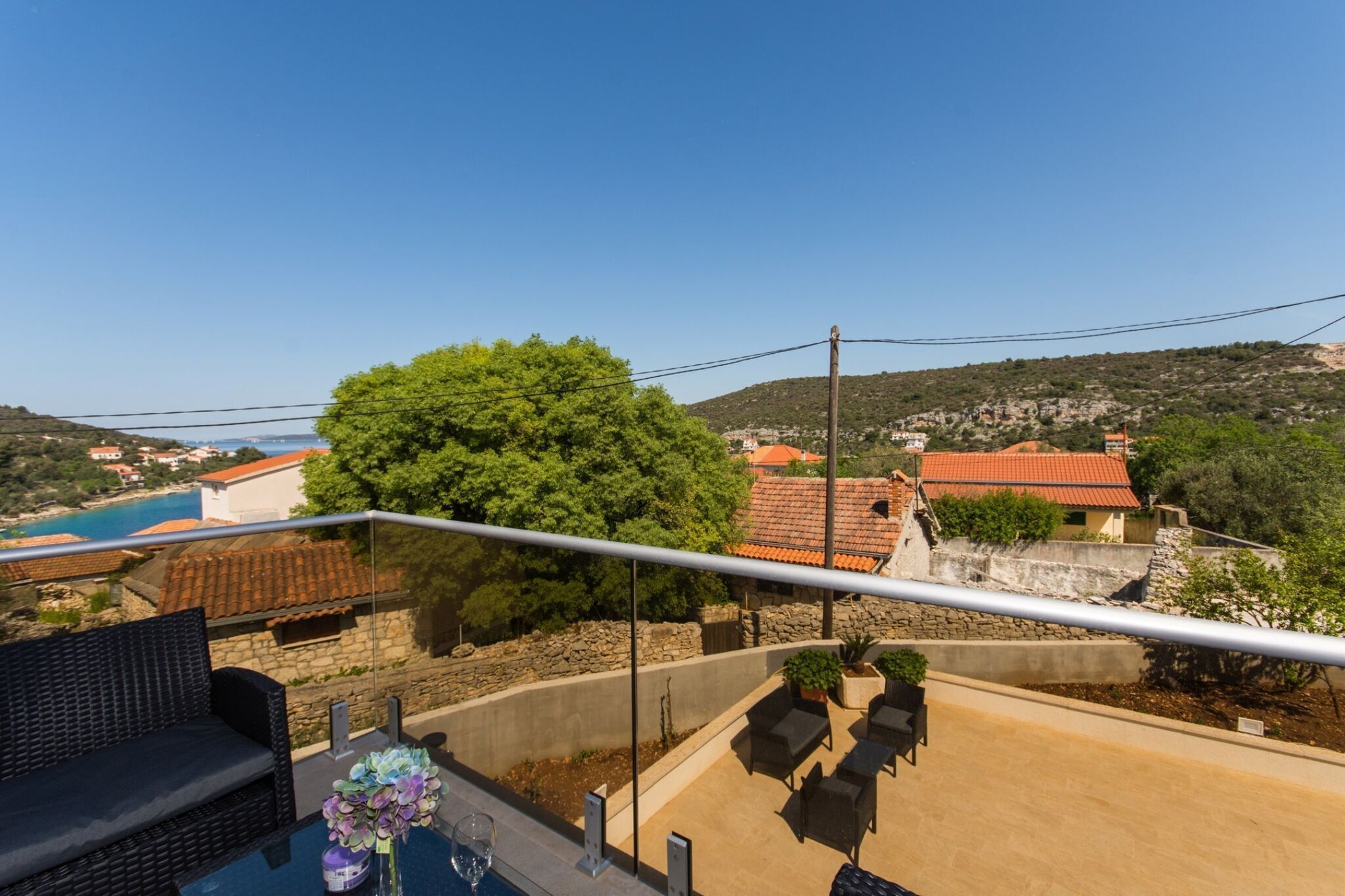 Un balcone con mobili da esterno, vista su una cittadina costiera e un albero di grandi dimensioni.