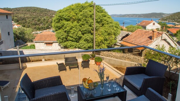 A balcony with wicker furniture, a glass table with fruit and drinks, and a view of a coastal town and hills.