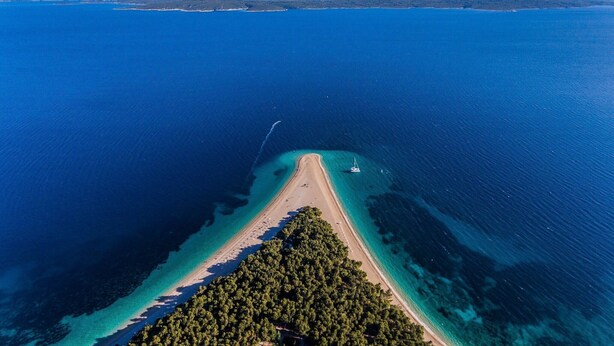 An aerial view of a coastal area with a beach, clear waters, and a forested peninsula.