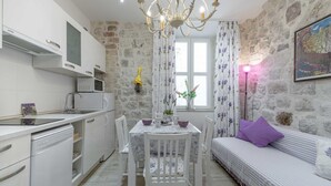 A compact kitchen with white cabinetry, a built-in microwave, and a dishwasher. A dining table with chairs is set against a stone wall, and a sofa with a purple cushion is visible in the background.