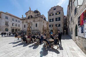 Un café al aire libre con mesas y sillas, rodeado de edificios históricos con fachadas de piedra y ventanas arqueadas.