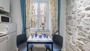 A compact kitchen with a microwave and a small refrigerator, a dining table with chairs, and a window with curtains. - L & L Old Town Apartments - Studio Apartment ( Lacroma) (Dubrovnik)