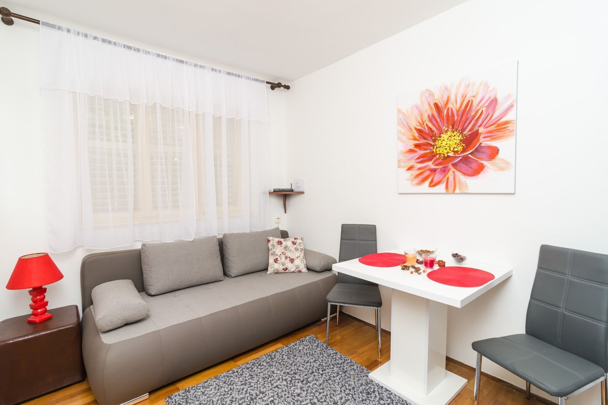 Un salon moderne avec un canapé gris, une petite table à manger agrémentée de sets de table rouges et une lampe rouge posée sur une table d’appoint en bois.