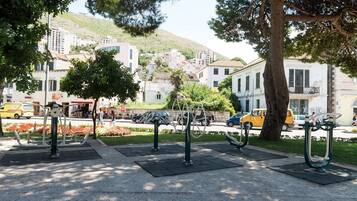 Un parc équipé d’installations d’exercice en plein air, doté d’une zone dallée et offrant en toile de fond un paysage urbain avec des bâtiments et une colline.