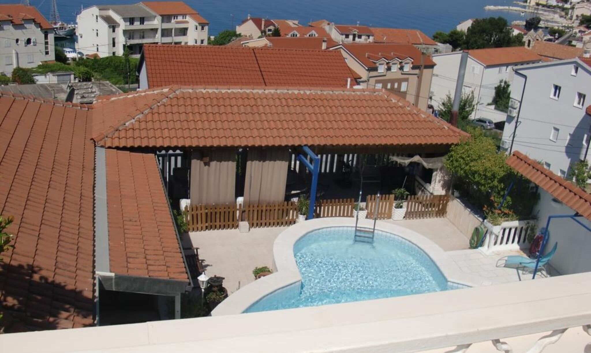 A residential area with a swimming pool, red-tiled roofs, and a view of the sea.