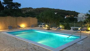 Une piscine rectangulaire bordée d’une terrasse en pierre, entourée de chaises longues et d’une clôture en chaume.