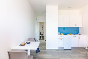 Uma cozinha moderna com armários brancos e painel azul, uma área de refeições com mesa e cadeiras brancas, e uma porta visível que dá para outro cômodo.