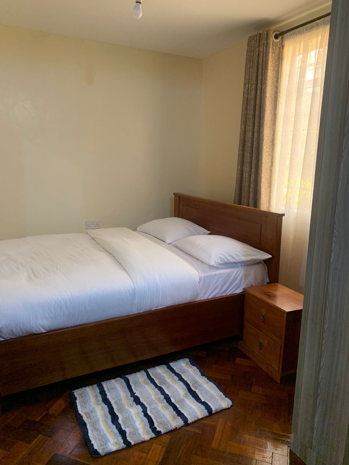 A single bed with white bedding, a wooden headboard, a nightstand, and a striped rug on a wooden floor.