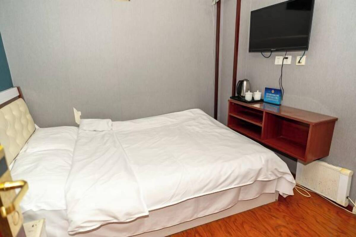 Una cama individual con ropa de cama blanca, un televisor montado en la pared y un estante de madera con una tetera y tazas.